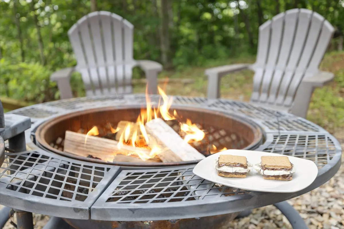 A cozy fire pit surrounded by Adirondack chairs is the focal point. S'mores supplies rest nearby, and the soft glow of the fire is reflected by lush greenery in the background, evoking a sense of warmth and togetherness in nature.