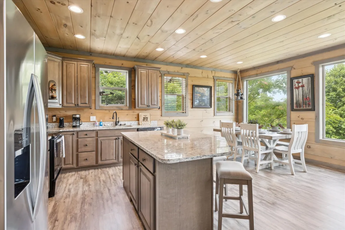 A warmly lit kitchen and dining space featuring a granite countertop, wooden cabinets, and a dining area with white chairs. Large windows offer lush greenery views, filling the space with natural light. The setup invites group cooking and shared meals in a cheerful environment.