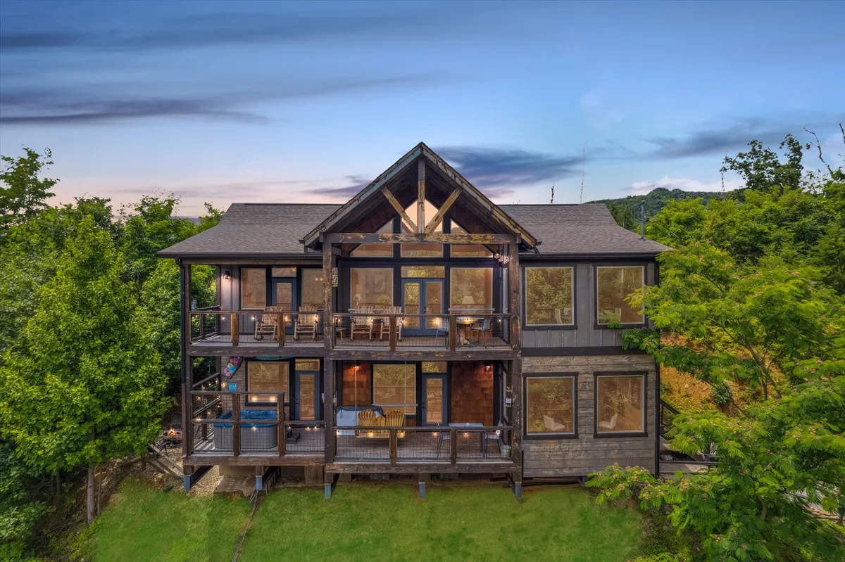 The image showcases the exterior of a modern cabin nestled in lush greenery, with expansive decks and balconies offering ample space for outdoor dining and relaxation. Large windows allow natural light to flood the interior, and the sunset can be seen casting a warm glow over the spacious deck, which is perfect for gatherings, enjoying morning coffee, or stargazing in the evening.