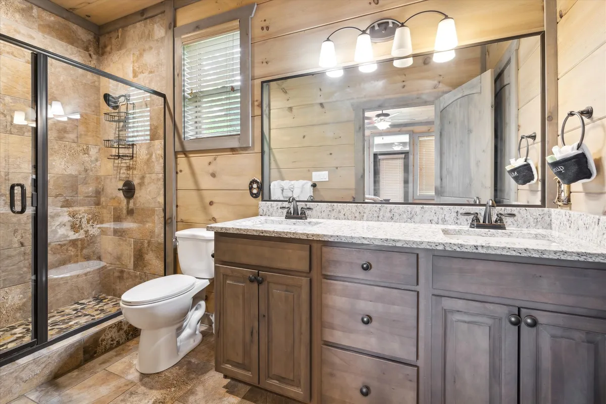 A modern, tranquil bathroom with double sink vanity, granite countertop, glass shower enclosure, and rustic wood paneling. The spacious layout and calming mood make it an ideal space for self-care and relaxation.