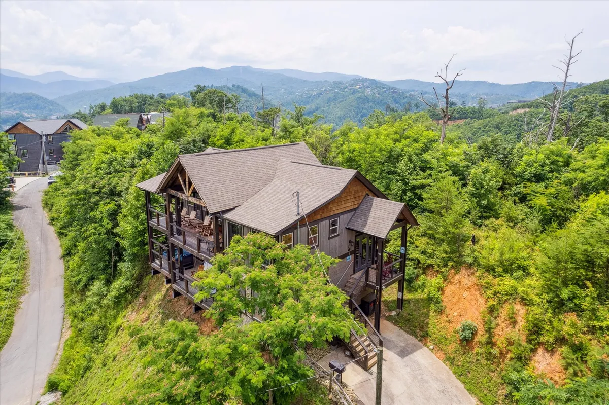 A broad exterior shot of the house with expansive windows and a mountain backdrop, surrounded by greenery. The porch and setting evoke the gateway to both nature and nearby Gatlinburg attractions.