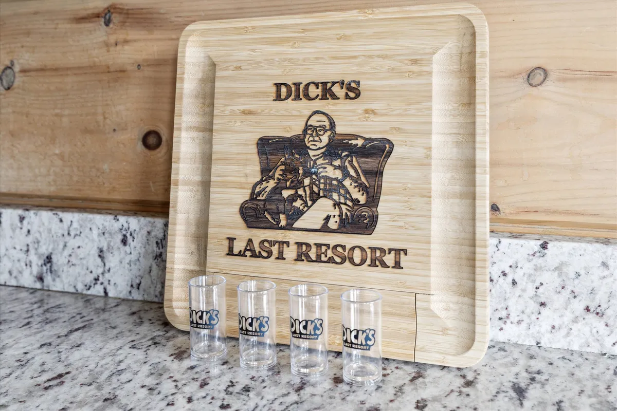A close-up of a wooden tray engraved with a playful message, adorned with shot glasses ready for drinks, inviting friends to celebrate together. The setting suggests a casual, festive atmosphere ideal for pre-nightlife gatherings or moonshine tastings.