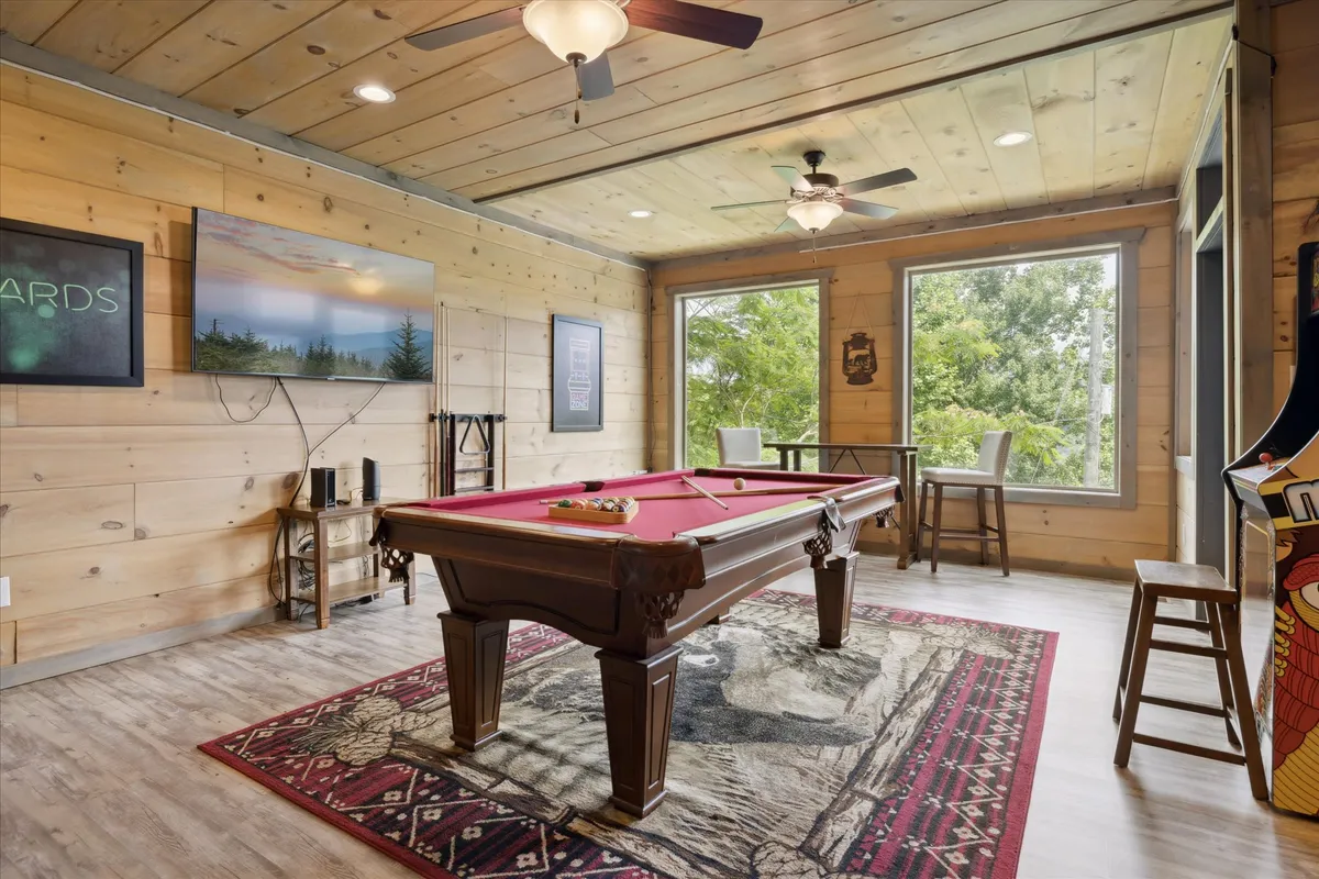A spacious entertainment lounge with a pool table, large windows framing the view, wooden walls for rustic charm, a large television set, and bar stools. The layout encourages both game play and relaxed viewing or conversation.