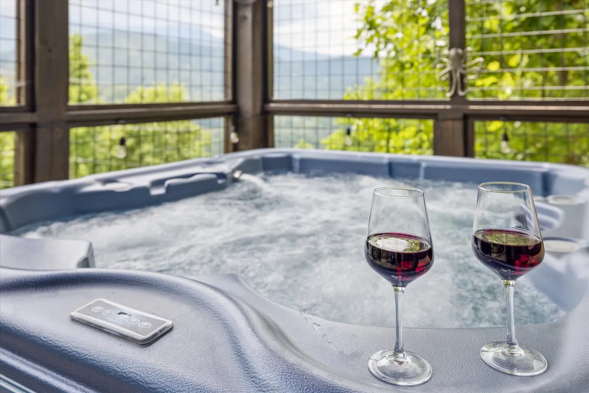 A bubbling hot tub sits on the deck, flanked by two wine glasses resting on the edge. In the background, a sweeping view of the Smoky Mountains is visible beneath an evening sky, suggesting a perfect setting for relaxation or stargazing.