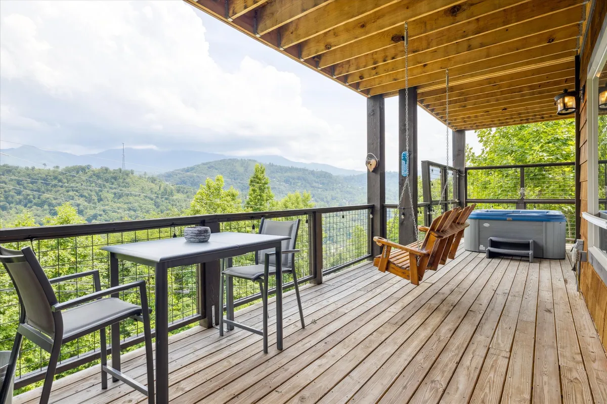 An inviting deck features a hot tub, outdoor seating, and a dining table. The scene overlooks a panoramic mountain view, blending luxury and natural beauty, with the setup hinting at al fresco dining or relaxing after a soak.