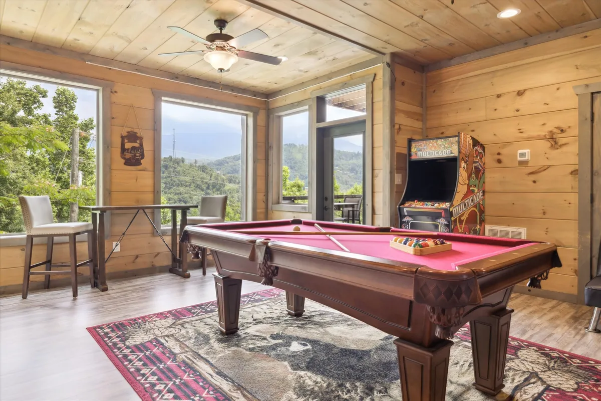 A bright and welcoming game room featuring a modern pool table at the center, classic arcade machine, wooden floors, and expansive windows that let in natural light. The scene captures people playing pool and arcade games while others socialize and enjoy the sunset views outside.