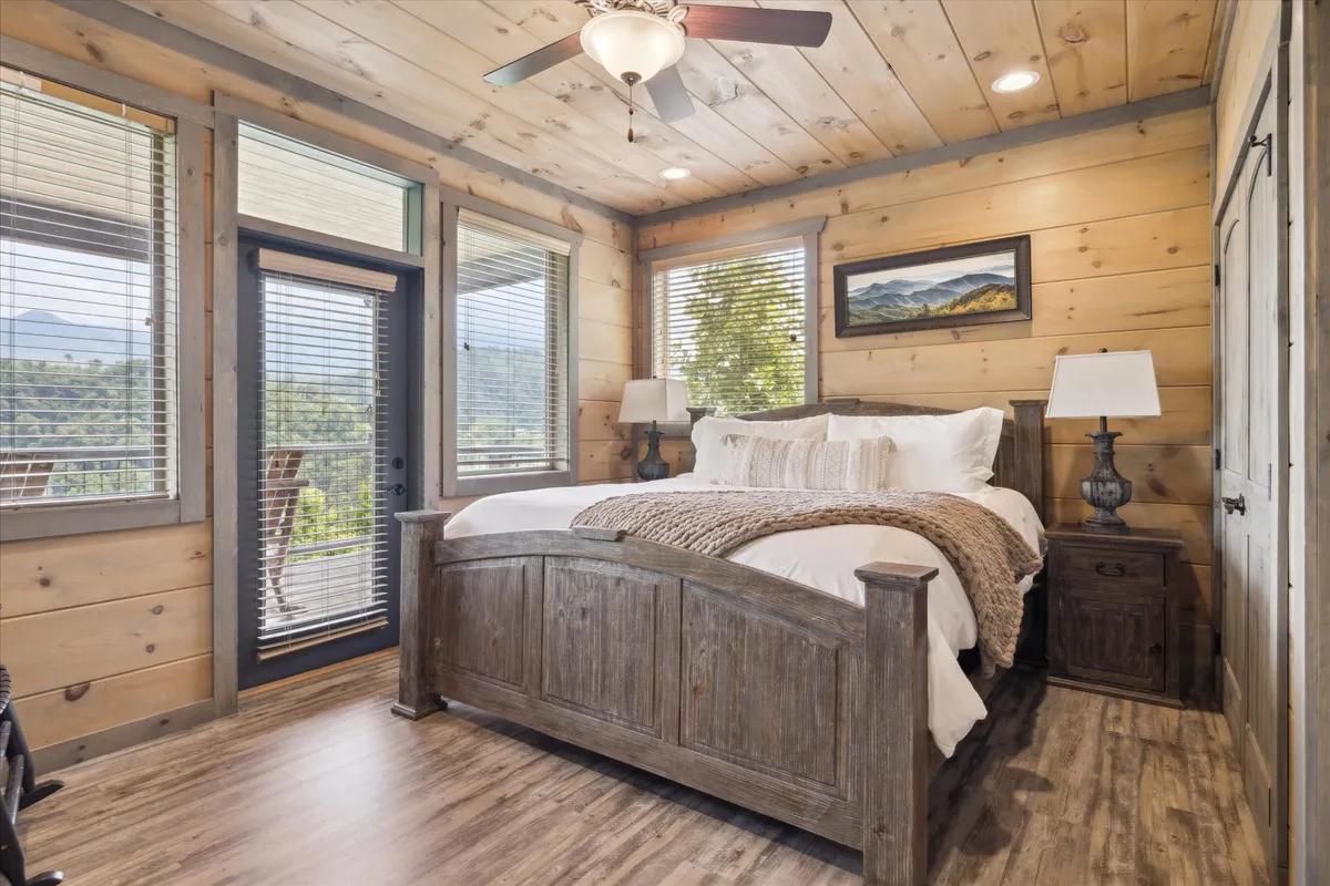 A serene bedroom featuring a large wooden bed with plush bedding, a nightstand with a lamp, and expansive windows that invite natural light. A mountain view painting above the bed and soft lighting contribute to a peaceful, inviting atmosphere. The overall setting encourages relaxation and restful sleep.