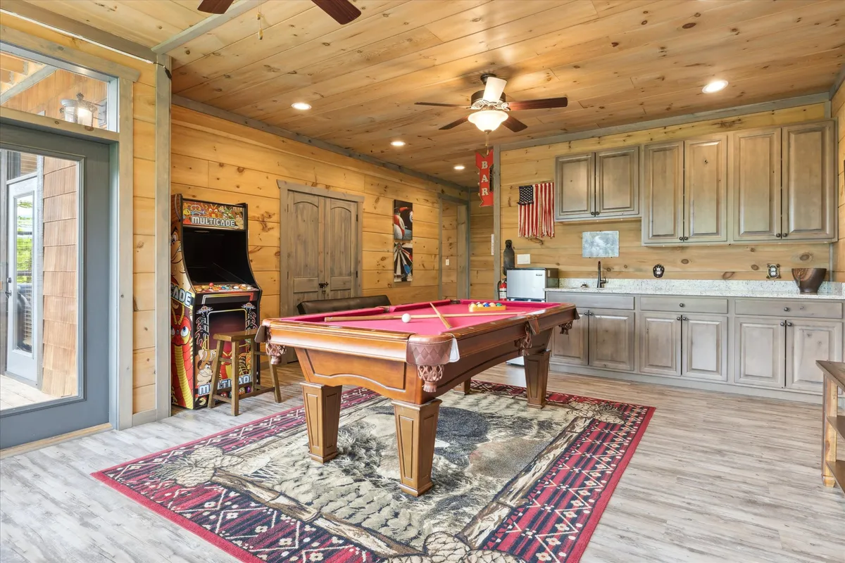 A cozy game retreat with a pool table in the foreground, an arcade game machine, bar area, and warm wooden accent walls. Friends can be seen enjoying drinks and friendly competition.