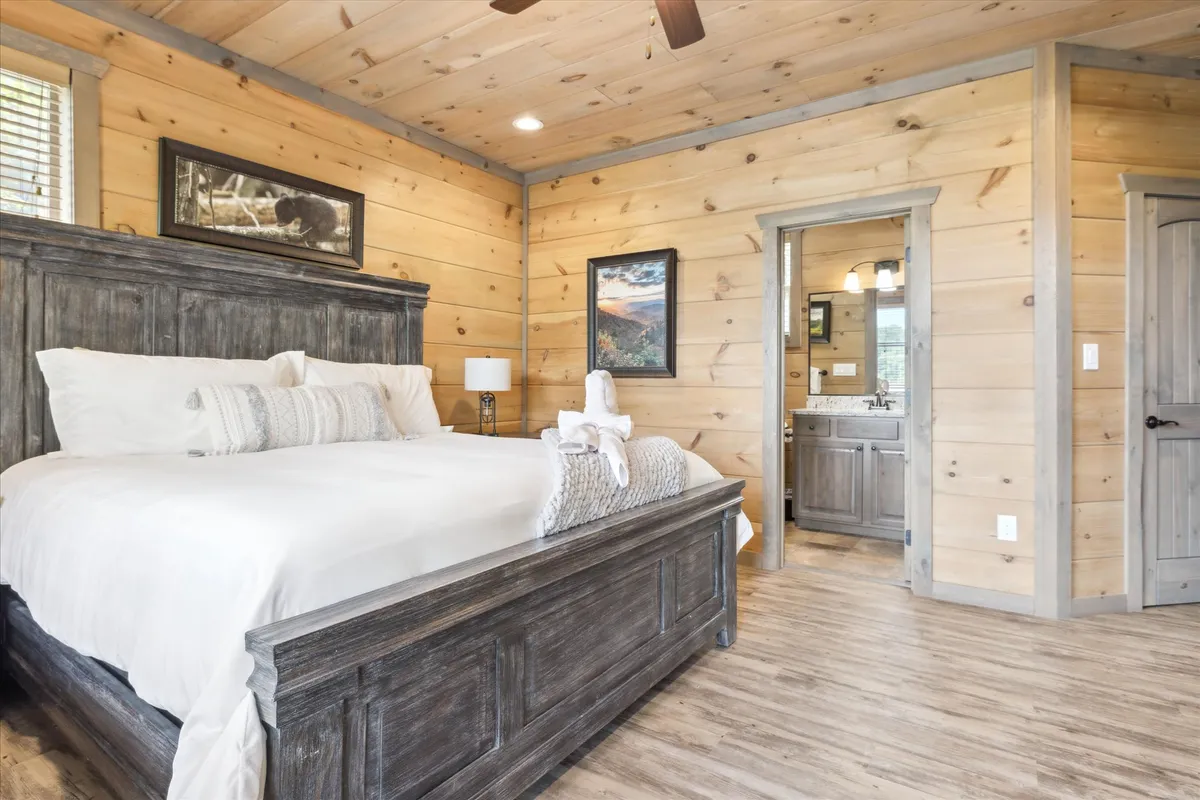 A relaxing bedroom with a large wooden bed adorned with decorative pillows and fluffy blankets. The room features artwork on the walls and a visible bathroom entrance, hinting at ensuite convenience. Soft lighting and neutral tones evoke comfort and serenity.