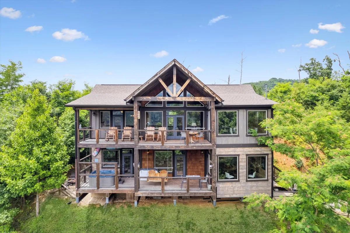 A spacious balcony featuring wooden rocking chairs and a large wooden table, surrounded by lush green foliage and expansive windows that reflect the outdoors. The scene conveys a welcoming outdoor space ideal for gatherings, relaxation, or winding down after a night out.