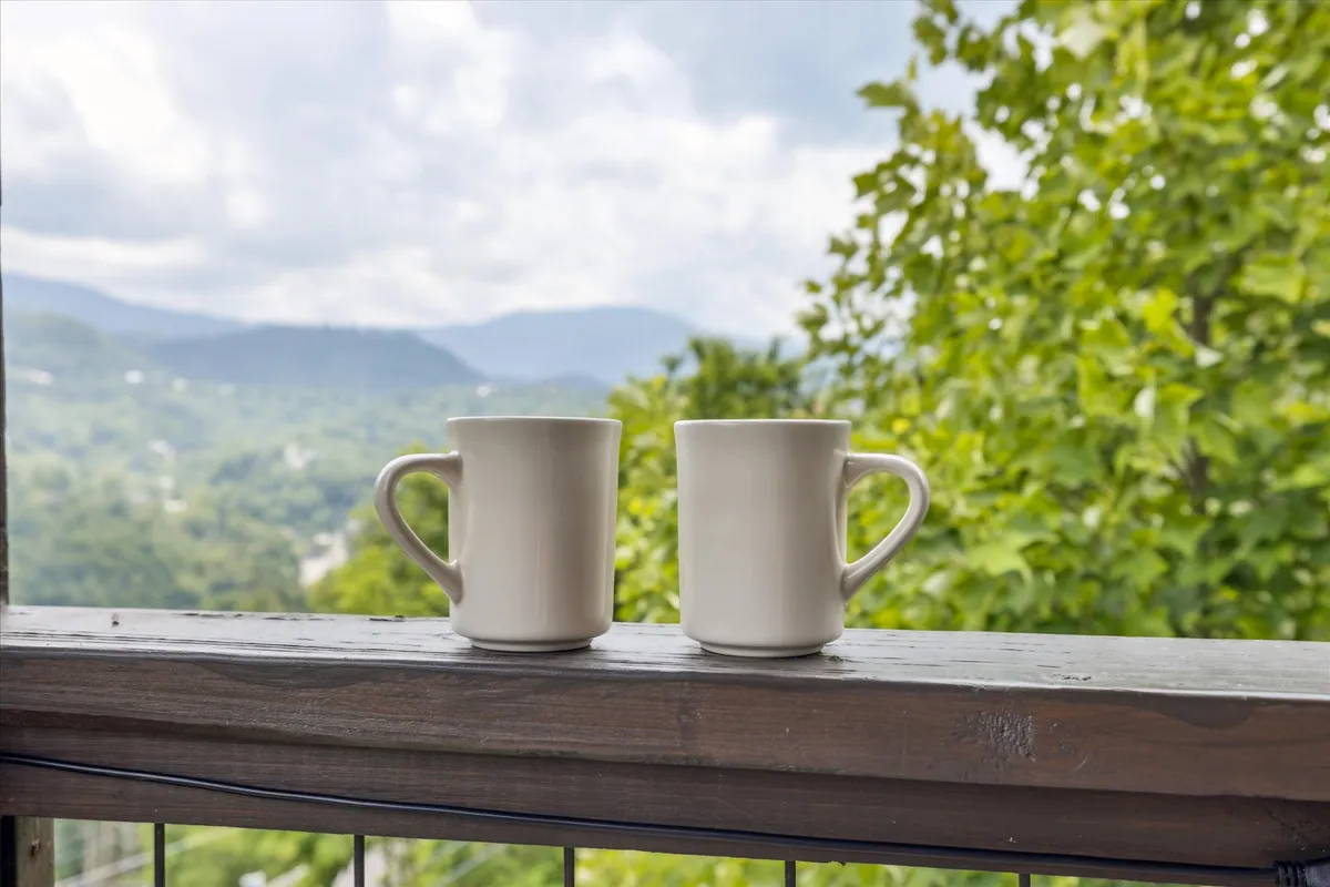 This image features two white mugs resting on a balcony with lush green trees and a sweeping mountain view in the background. The outdoor setting invites viewers to imagine leisurely mornings enjoying a warm drink while taking in the natural beauty of the Smoky Mountains.